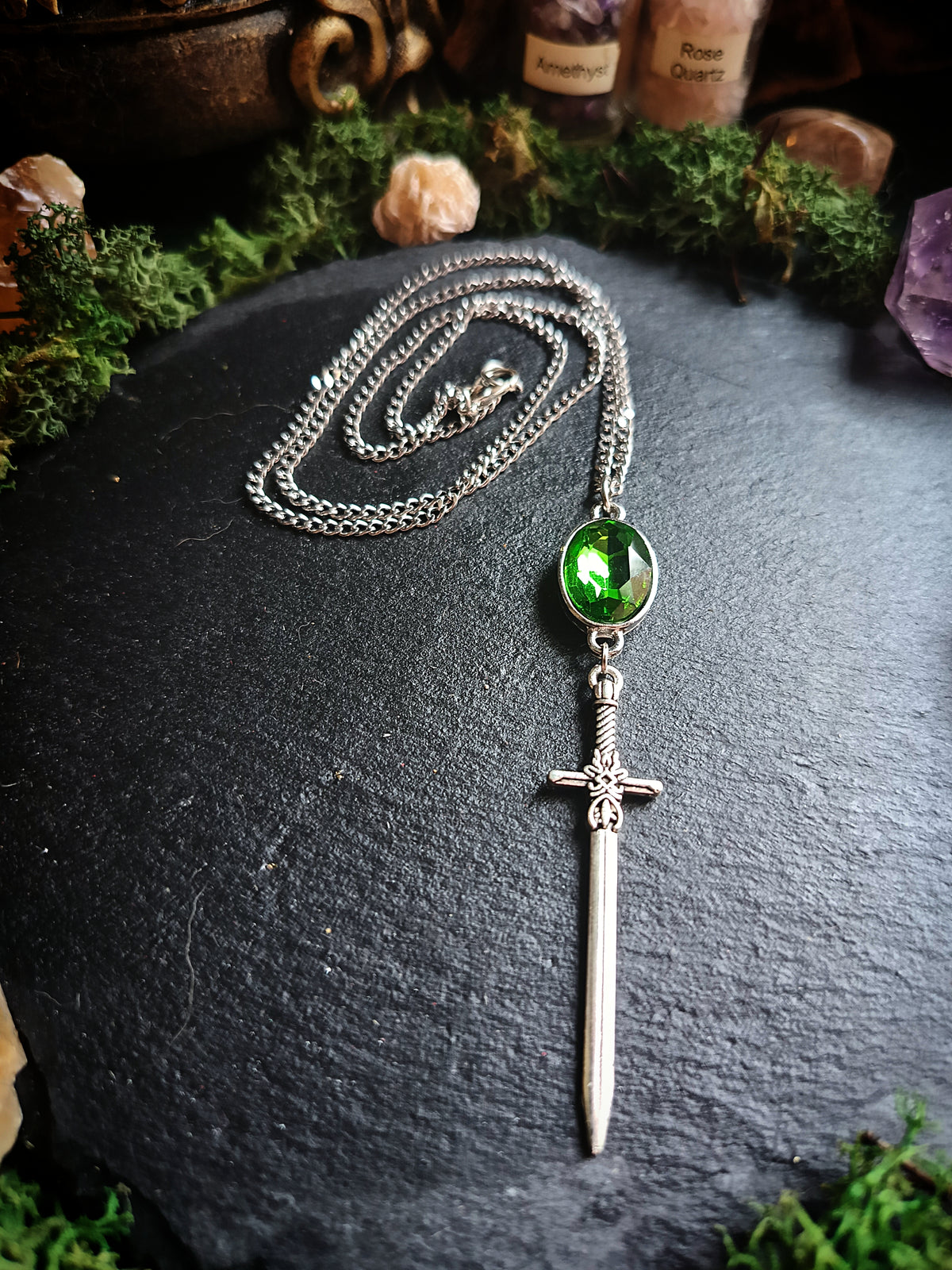 A silver-toned chain necklace featuring a round green gemstone and a detailed sword pendant, displayed on a dark slate surface with moss and crystals in the background.
